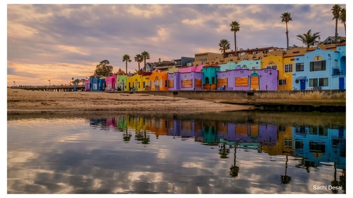 Colorful houses line the beach. Palm trees stand behind them. Their reflection can be seen in the still water nearby.